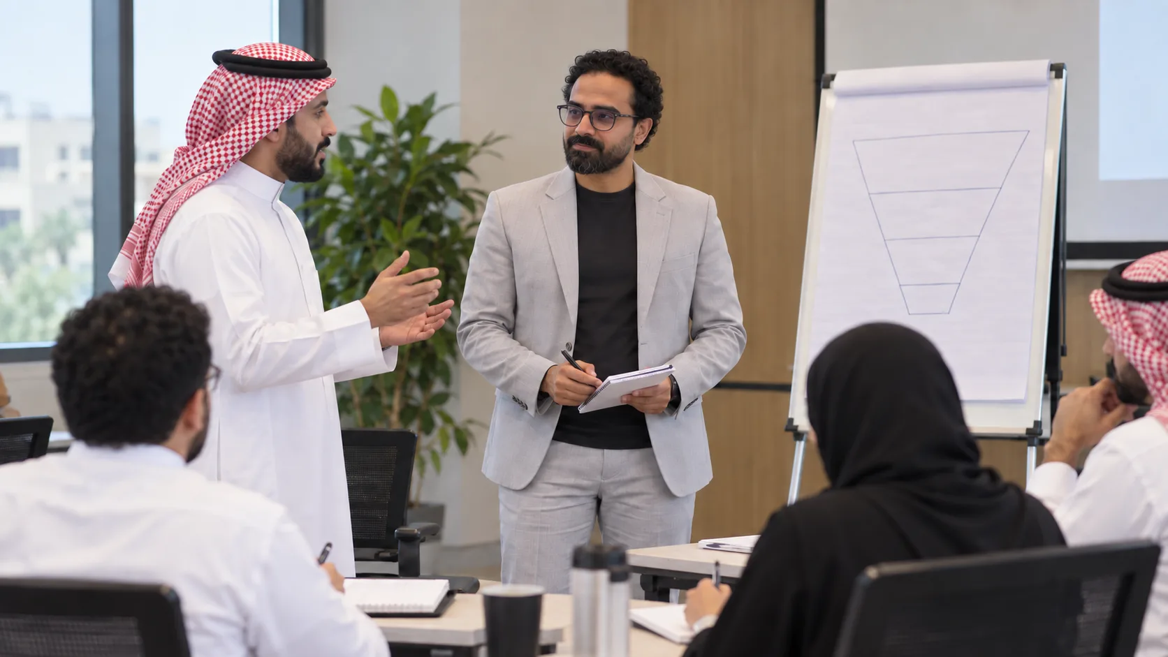 Ashraf Ibrahim beside a flip chart with a sales-funnel sketch, listening to a standing participant while a small group around a training table follows along